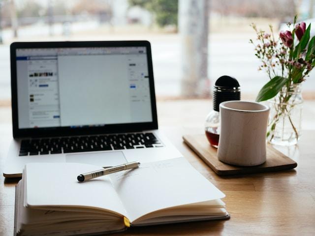 A laptop over a desk with a notebook, pen, and a cup of tea with a flower vase